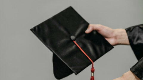 Image of two lighter skinned hands holding a black graduation cap with a red tassel and a white diploma with a red ribbon.