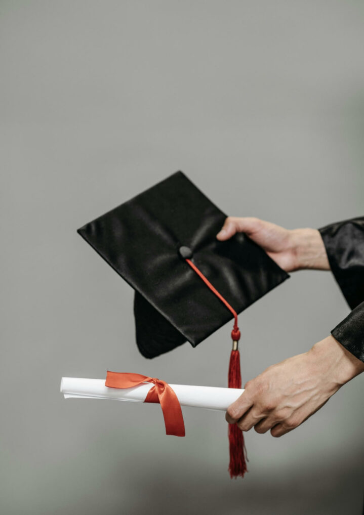 Image of two lighter skinned hands holding a black graduation cap with a red tassel and a white diploma with a red ribbon.