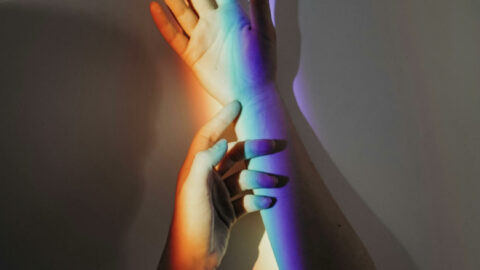 Two arms being held up in the air. One hand is touching the other hand by the wrist. There is a rainbow shadowing over both hands.