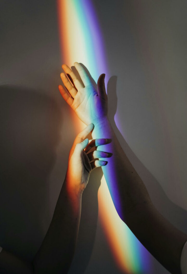 Two arms being held up in the air. One hand is touching the other hand by the wrist. There is a rainbow shadowing over both hands.