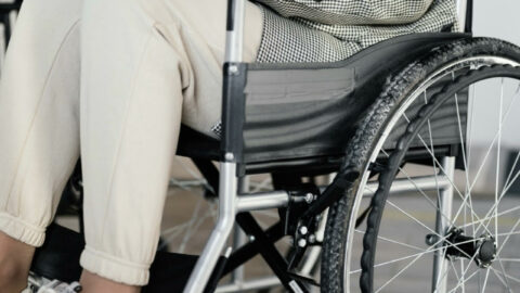 Image of a persons body sitting in a wheelchair working on their laptop. This person is wearing a white hoodie, black and white blazer over top, cream colored joggers and white sneakers.