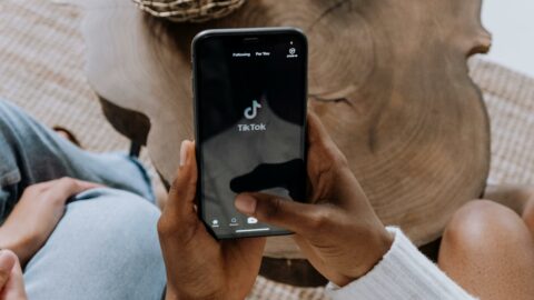 2 darker skinned hands holding an iPhone that is opened to the TikTok app. There is a brow wooden coffee table in the background.