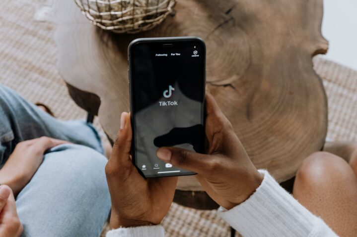 2 darker skinned hands holding an iPhone that is opened to the TikTok app. There is a brow wooden coffee table in the background.