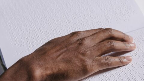 This image shows a close-up of a hand reading Braille, a tactile writing system used by people who are visually impaired. The hand is gently placed on a page filled with raised dots, which form Braille characters. The person’s skin tone is medium-dark, and they are wearing a light blue sleeve. The white page contrasts with the hand, highlighting the texture of the Braille script