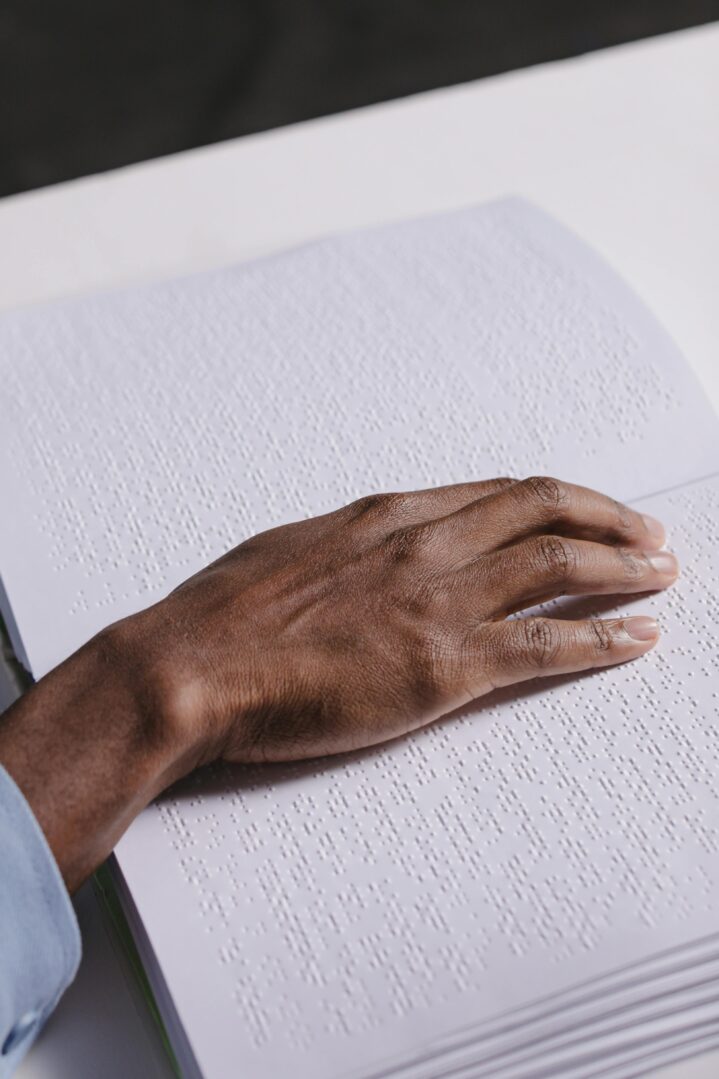 This image shows a close-up of a hand reading Braille, a tactile writing system used by people who are visually impaired. The hand is gently placed on a page filled with raised dots, which form Braille characters. The person’s skin tone is medium-dark, and they are wearing a light blue sleeve. The white page contrasts with the hand, highlighting the texture of the Braille script