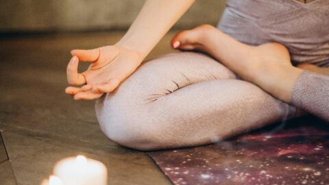 The image shows a person sitting cross-legged on a yoga mat, meditating. They are wearing light-colored leggings and a white sports bra, with one hand resting on their knee in a traditional meditation pose. In the foreground, there are lit candles, creating a warm and calming atmosphere. The scene evokes a sense of peace and mindfulness.