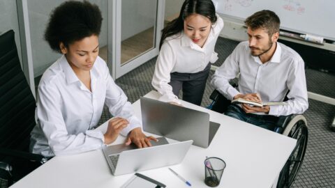 The image shows three people gathered around a desk, working together on laptops. One person, a man using a wheelchair, is taking notes in a notebook while watching the others. Another person, seated, is typing on a laptop and wearing a focused expression. The third person is standing, leaning over slightly to engage with the seated team members. Behind them, there is a whiteboard with various charts and notes, and the setting appears to be a modern office space, suggesting a collaborative and inclusive work environment.