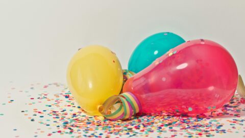 3 balloons, one pink, one yellow, and one blue, are laying on a bed of rainbow confetti.