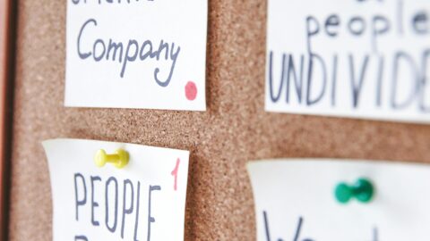 A corkboard with several handwritten notes pinned to it using colorful thumbtacks. The notes feature motivational and people-focused phrases, such as "HUMAN-oriented Company" written in black and blue ink with a red dot in the bottom right corner. Another note below it reads "PEOPLE FIRST" in bold black letters with a similar red dot. To the right, partially visible notes say "business & people UNDIVIDED" and "We start from WHY." The overall composition conveys themes of teamwork, human-centered values, and collaboration.