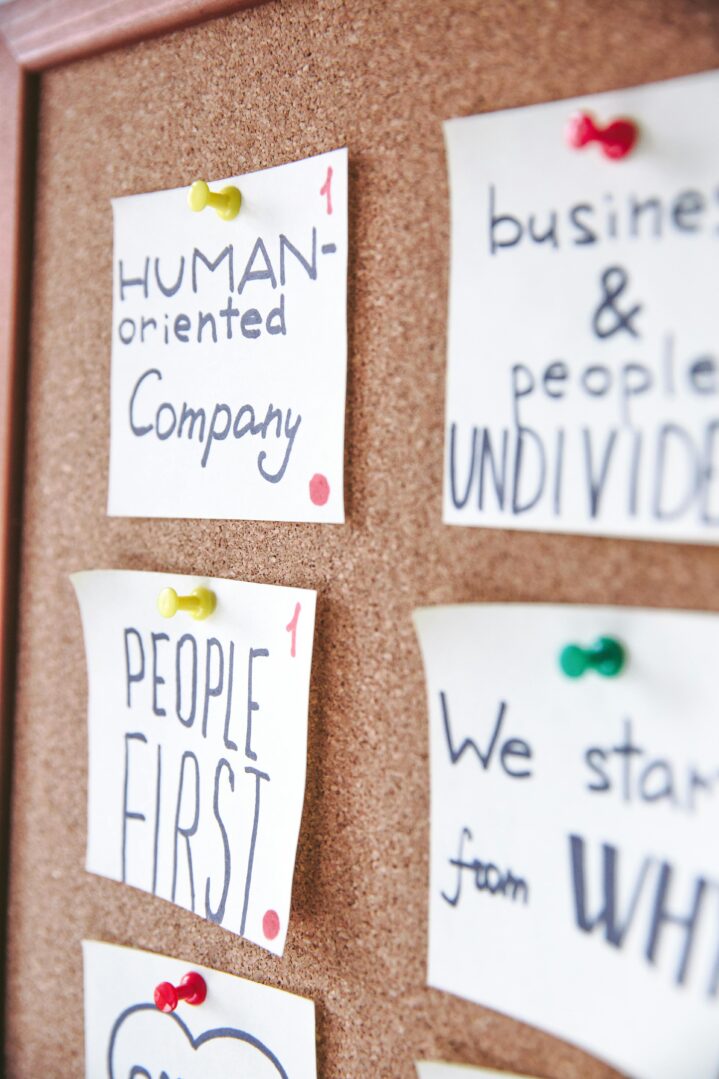 A corkboard with several handwritten notes pinned to it using colorful thumbtacks. The notes feature motivational and people-focused phrases, such as "HUMAN-oriented Company" written in black and blue ink with a red dot in the bottom right corner. Another note below it reads "PEOPLE FIRST" in bold black letters with a similar red dot. To the right, partially visible notes say "business & people UNDIVIDED" and "We start from WHY." The overall composition conveys themes of teamwork, human-centered values, and collaboration.